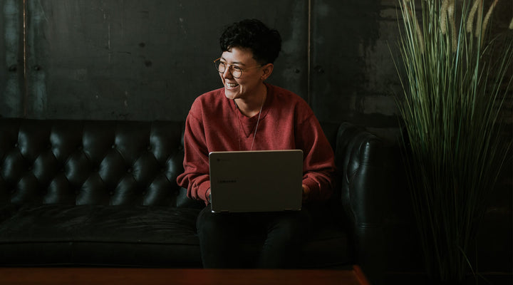 Smiling individual working on a laptop while seated on a stylish black leather couch, embodying creativity and productivity in a relaxed, modern environment