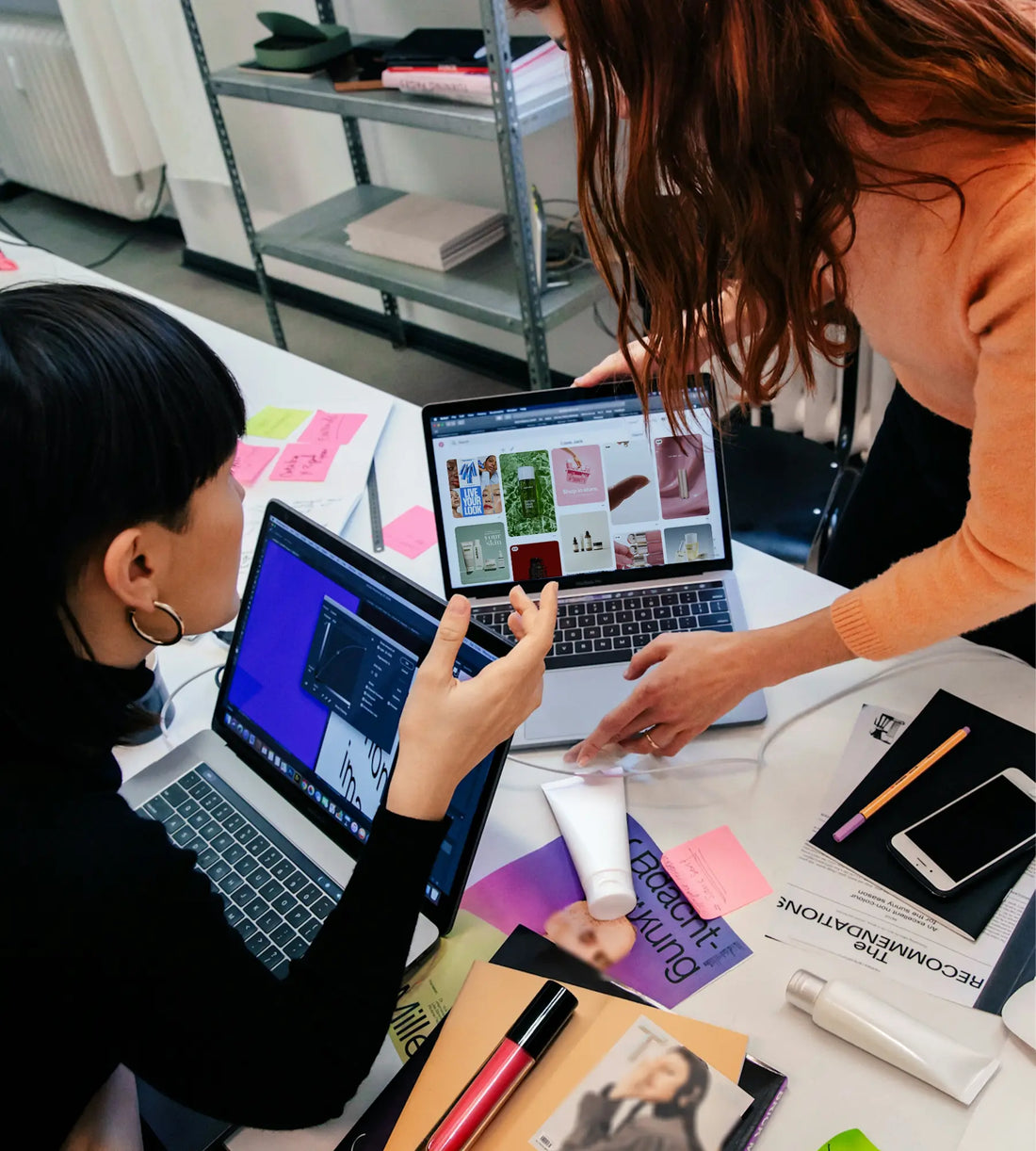 Two women collaborate on beauty product branding at a desk with laptops, packaging samples, and design mockups.