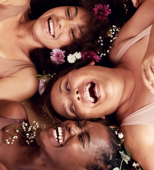 Three women with natural hair and flowers in their curls, lying together and laughing. A joyful, inclusive moment celebrating beauty and community.