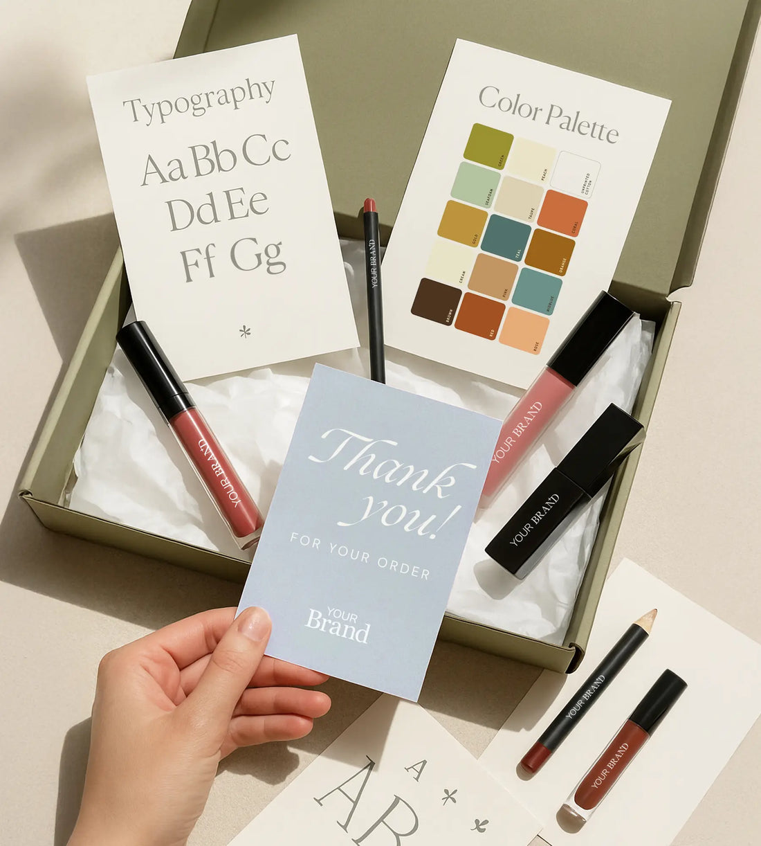 A hand holding a branded thank you card above a beauty packaging box with lipsticks, lip liners, and design reference cards for typography and color palette.