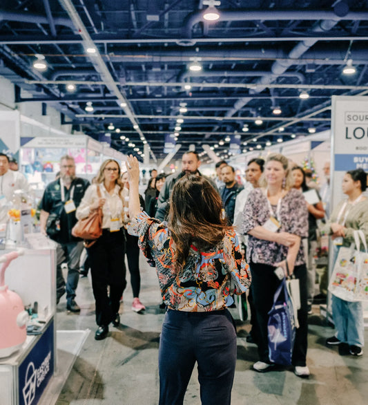 A woman speaking to a crowd at a busy trade show, with multiple booths and attendees in the background.