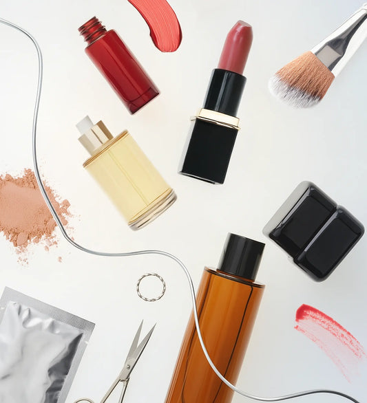 Overhead flatlay of various beauty products including lipstick, serum bottles, a makeup brush, powder, scissors, and red swatches on a white background.