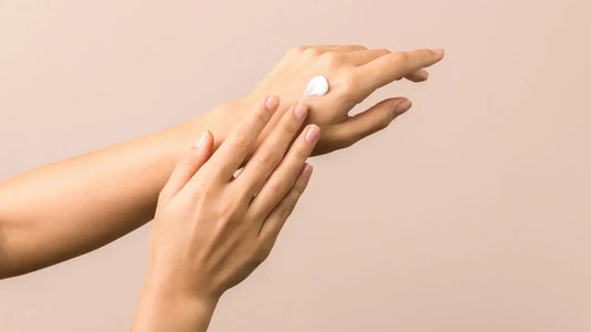 Close-up of hands applying a small dollop of cream to the back of one hand against a neutral beige background