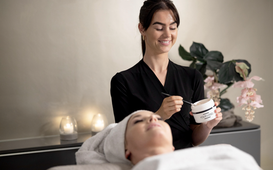 A medspa technician applying a mask on a woman's face lying down on the practitioner's bed with candles around