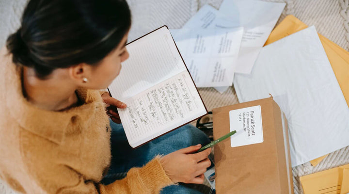 Overhead view of a woman reviewing notes while preparing a labeled shipping package, showcasing organization and best practices in e-commerce fulfillment