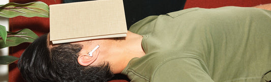 A guy laying down with a book covering his face and an airpod in ear wearing a green tshirt