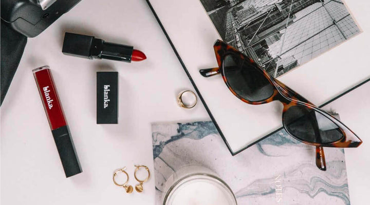 Flat lay of Blanka red lipstick and lip gloss paired with stylish accessories like sunglasses, gold jewelry, and a candle, illustrating sophistication and branding in the beauty industry