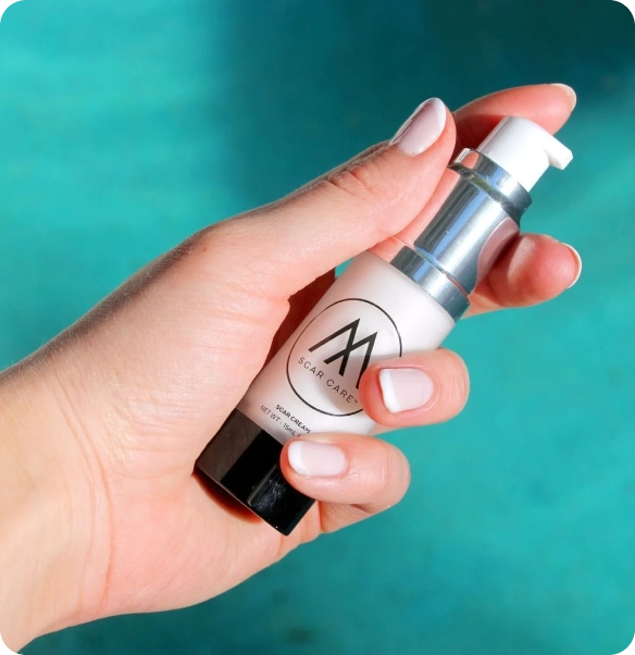 A hand with pale pink manicured nails holds a small skincare pump bottle against a bright turquoise background. The bottle is white with a black cap/base and a silver pump collar, featuring a circular logo with an 'M' and the text 'SCAR CARE'.