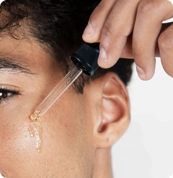 A close-up of a person's face and ear as a hand holds a clear dropper above the skin, with a small amount of golden serum being applied.